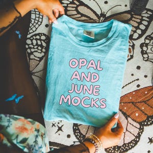 Peut inclure: Un t-shirt bleu clair avec le texte "OPAL AND JUNE MOCKS" imprimé en lettres roses.