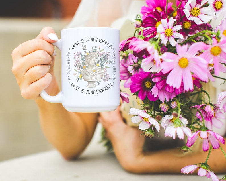Puede incluir: Taza de cer&aacute;mica blanca con un dise&ntilde;o floral y el texto "OPAL & JUNE MOCKUPS." La taza est&aacute; siendo sostenida por una mano.