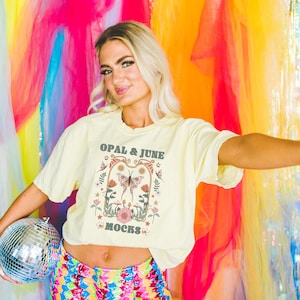 May include: A woman wearing a yellow t-shirt with a butterfly and floral design and the text "OPAL & JUNE MOCKS". She is holding a silver disco ball in her left hand and her right arm is extended out to the side.