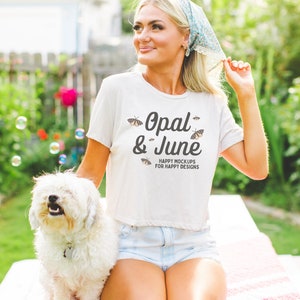 May include: A woman wearing a white t-shirt with the text "Opal & June" and butterflies printed on it. She is sitting on a pink and white blanket with a white dog beside her. The text below the logo reads "Happy Mockups for Happy Designs".