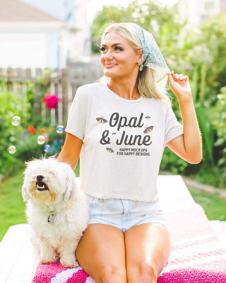 May include: A woman wearing a white t-shirt with the text "Opal & June" and butterflies printed on it. She is sitting on a pink and white blanket with a white dog beside her. The text below the logo reads "Happy Mockups for Happy Designs".