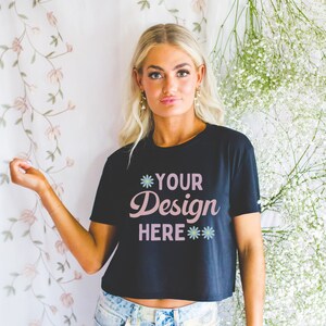Może przedstawiać: Czarny, kr&oacute;tki T-shirt z napisem "*YOUR Design HERE**" w r&oacute;żowych literach. Koszulkę nosi osoba z blond włosami, stojąca przed białym tłem z białymi kwiatami.
