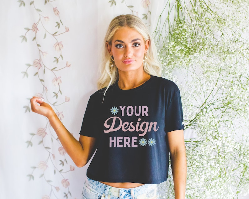 Może przedstawiać: Czarny, kr&oacute;tki T-shirt z napisem "*YOUR Design HERE**" w r&oacute;żowych literach. Koszulkę nosi osoba z blond włosami, stojąca przed białym tłem z białymi kwiatami.