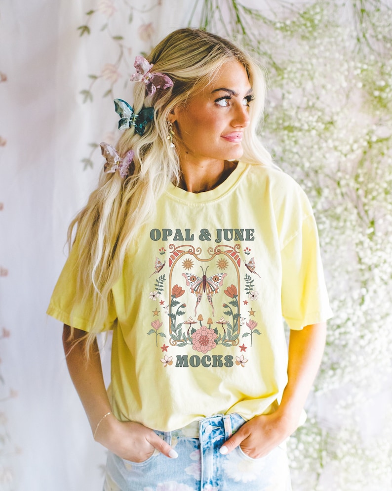 Pu&ograve; includere: Una maglietta gialla con un design grafico che presenta una farfalla, fiori e il testo "OPAL & JUNE MOCKS".