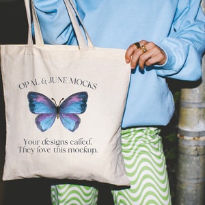 以下が含まれることがあります： 「OPAL & JUNE MOCKS」という文字と、紫と青の蝶のグラフィックが描かれた白いキャンバス地のトートバッグ。蝶の下のテキストには、「Your designs called. They love this mockup.」と書かれています。