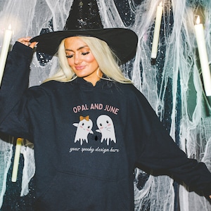 May include: Black hooded sweatshirt with the text "OPAL AND JUNE" and a graphic of two ghosts holding hands with a spider between them. The text "your spooky design here" is below the ghosts.