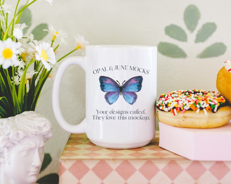 Pu&ograve; includere: Tazza in ceramica bianca con un motivo a farfalla blu e il testo "OPAL & JUNE MOCKS Your designs called. They love this mockup."