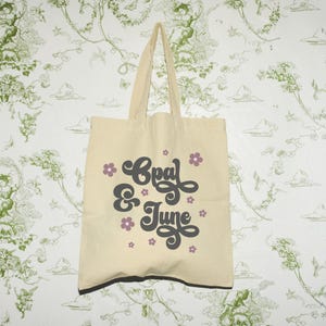 Könnte beinhalten: Beige Canvas-Tragetasche mit schwarzem Besatz und Griffen. Die Tasche zeigt den Text "Coal & June" in einer dunkelgrauen, stilisierten Schriftart mit kleinen lila Blumenakzenten. Der Hintergrund ist eine weiße Tapete mit einem grünen Blumenmuster.