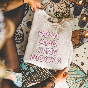 Puede incluir: Una sudadera de color crema con el texto "OPAL AND JUNE MOCKS" impreso en rosa en la parte delantera.