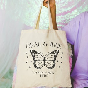 May include: A beige tote bag with black text that reads "OPAL & JUNE" and a black butterfly graphic. The text "YOUR DESIGN HERE" is below the butterfly.