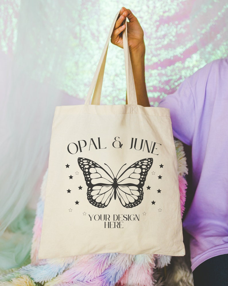 May include: A beige tote bag with black text that reads "OPAL & JUNE" and a black butterfly graphic. The text "YOUR DESIGN HERE" is below the butterfly.