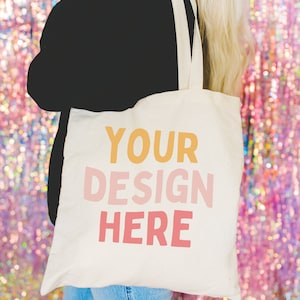 May include: A white tote bag with the text "YOUR DESIGN HERE" printed in yellow, pink, and orange. The bag is being held by a person wearing a black sweater and blue jeans.