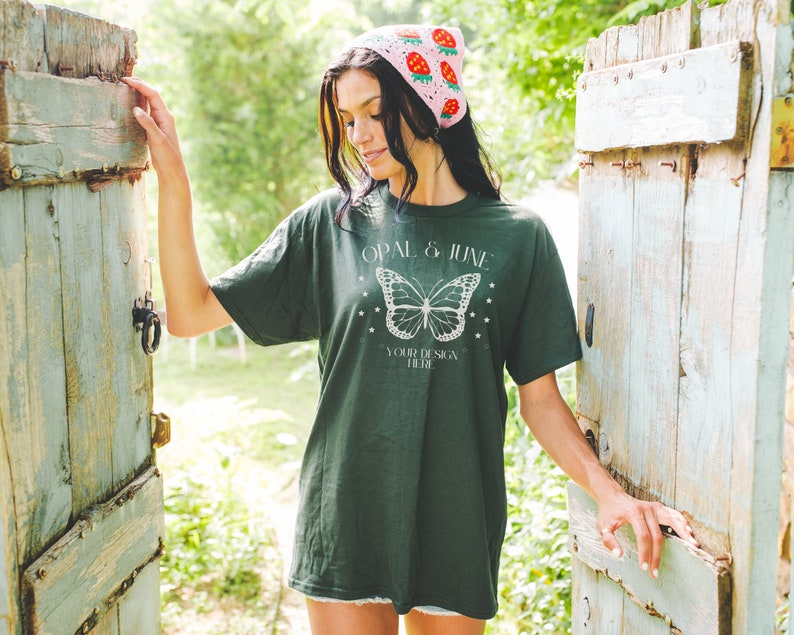 Pu&ograve; includere: Una donna indossa una maglietta verde con una grafica di farfalla bianca e il testo "Opal & June" e "Your Design Here". Indossa anche un cappello all'uncinetto rosa con disegni di fragole.