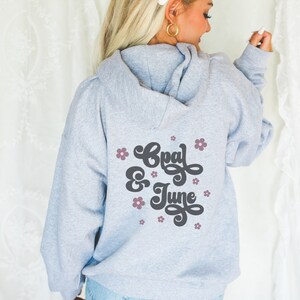 Puede incluir: Sudadera gris con capucha con el texto "Opal & June" en letra negra con flores rosas alrededor del texto.
