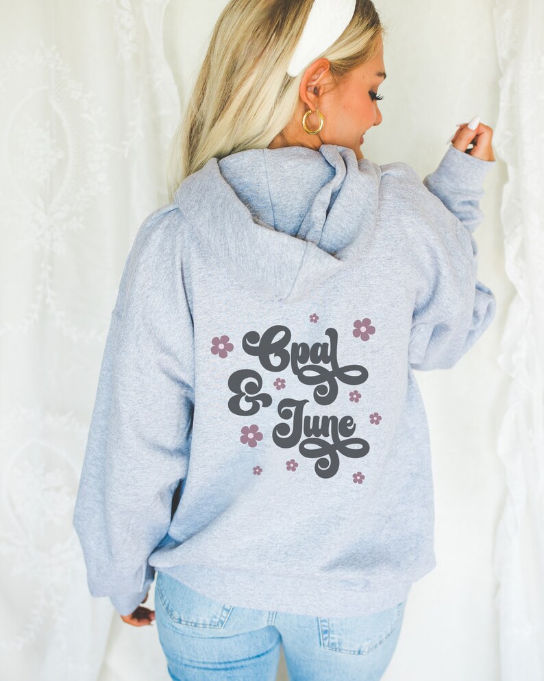 Puede incluir: Sudadera gris con capucha con el texto "Opal & June" en letra negra con flores rosas alrededor del texto.