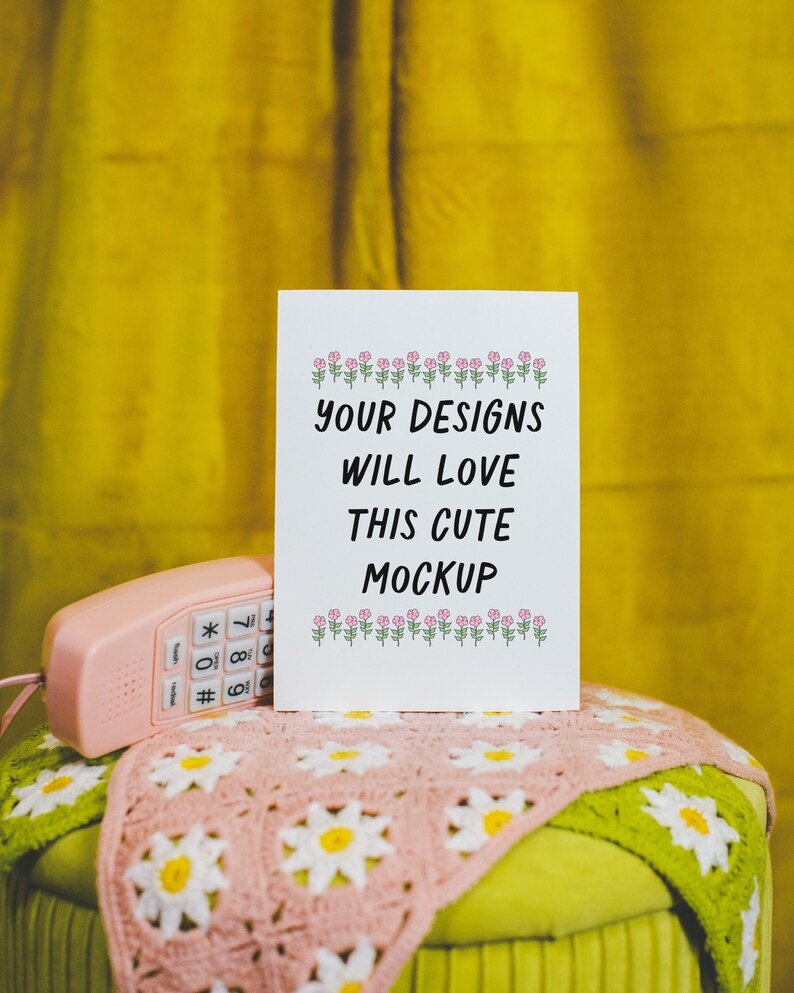 May include: A white card with the text "Your designs will love this cute mockup" and a row of pink flowers at the top and bottom. The card is sitting on a pink and green crocheted blanket.