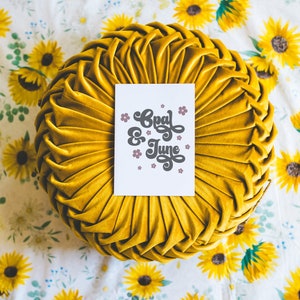 Op de afbeelding: Een rond kussen van geel fluweel met een witte kaart erop. De kaart heeft zwarte tekst die "Opal & June" zegt met kleine roze bloemen rond de tekst.