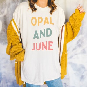 Puede incluir: Una camiseta blanca con las palabras "OPAL AND JUNE" impresas en letras de colores. La camiseta la lleva puesta una persona con el pelo largo y castaño y un cárdigan amarillo.