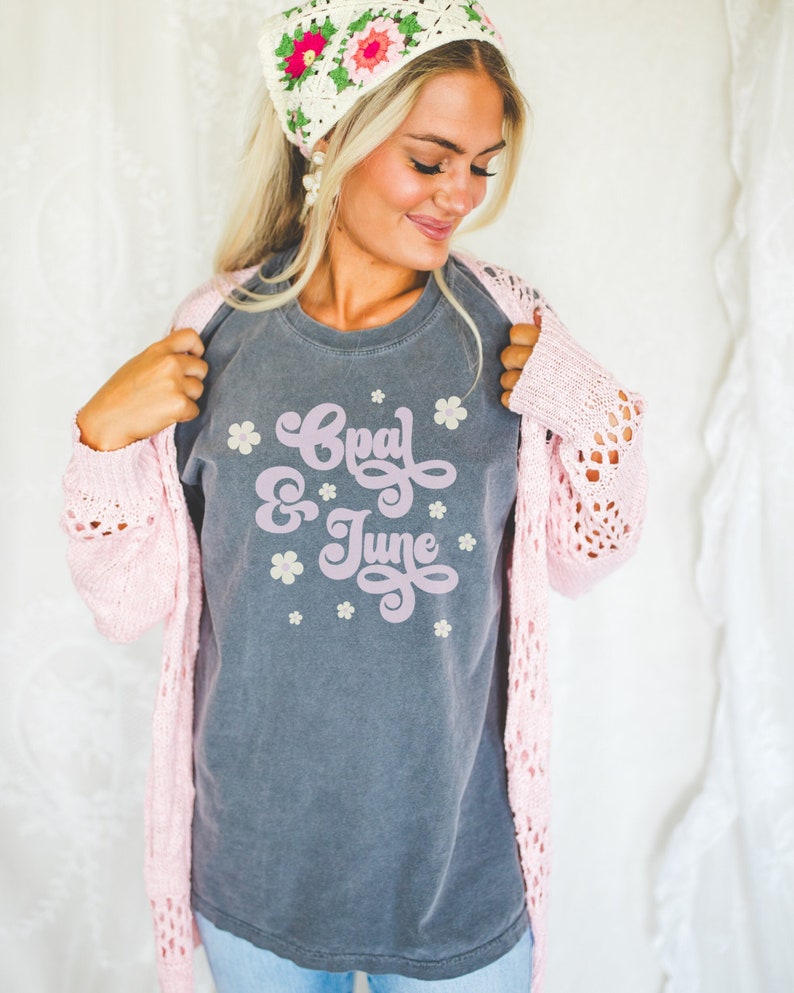 Puede incluir: Una camiseta gris con las palabras "Goal & June" en una fuente retro, rodeada de peque&ntilde;os gr&aacute;ficos de flores. La persona lleva una rebeca rosa y un pa&ntilde;uelo de ganchillo con detalles florales.