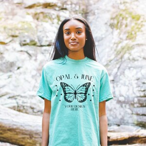 May include: A turquoise blue t-shirt with a black butterfly graphic and the text "Opal & June" and "Your Design Here".