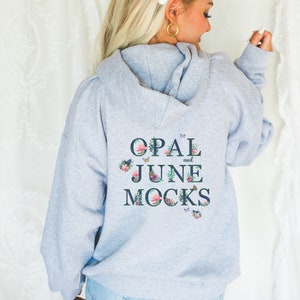 Puede incluir: Sudadera gris con capucha con el texto "OPAL and JUNE MOCKS" en letras azul oscuro con detalles florales y de mariposas.