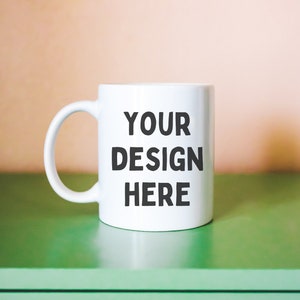 Puede incluir: Taza de cer&aacute;mica blanca con una inscripci&oacute;n negra que dice "YOUR DESIGN HERE" sobre una superficie verde.