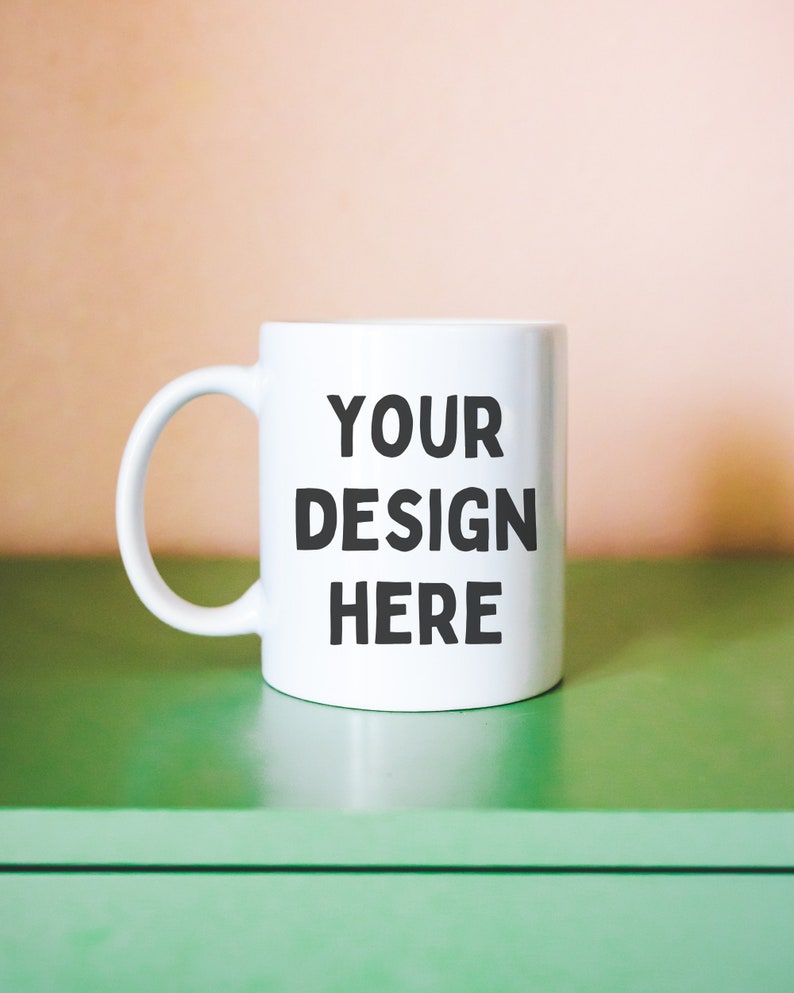 Puede incluir: Taza de cer&aacute;mica blanca con una inscripci&oacute;n negra que dice "YOUR DESIGN HERE" sobre una superficie verde.