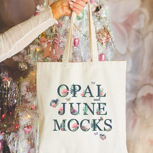 Puede incluir: Una bolsa de lona blanca con un diseño floral y el texto "OPAL and JUNE MOCKS" en letras azul oscuro.