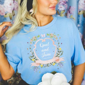 Könnte beinhalten: Ein hellblaues T-Shirt mit einem floralen Design und dem Text "Opal and June" auf einem rosafarbenen Banner. Das Design zeigt einen herzförmigen Rahmen mit einem Blumenkranz und Schmetterlingen.