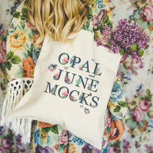 Può includere: Una borsa tote color crema con la scritta "OPAL and JUNE MOCKS" in un carattere blu scuro, decorata con elementi floreali e farfalle. La borsa è piena di un mazzo di fiori viola. La persona indossa una giacca a fantasia floreale.