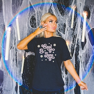 Puede incluir: Una mujer lleva una camiseta negra con el texto "Opal & June" en escritura rosa y detalles florales. La camiseta está sobre un fondo de red blanca y velas.