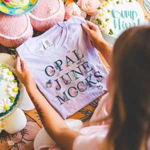 May include: A purple t-shirt with floral accents and the text "Opal and June Rocks" in dark blue lettering.