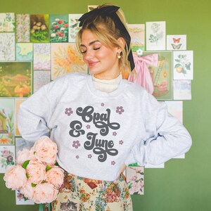 Puede incluir: Una sudadera gris con el texto "Opal & June" en una fuente retro con flores rosas alrededor del texto.