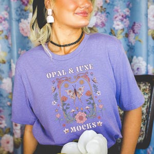 May include: A lilac-colored t-shirt with the text "OPAL & JUNE MOCKS" and a floral and butterfly graphic. The design includes butterflies, flowers, and decorative elements in shades of orange, pink, and green.