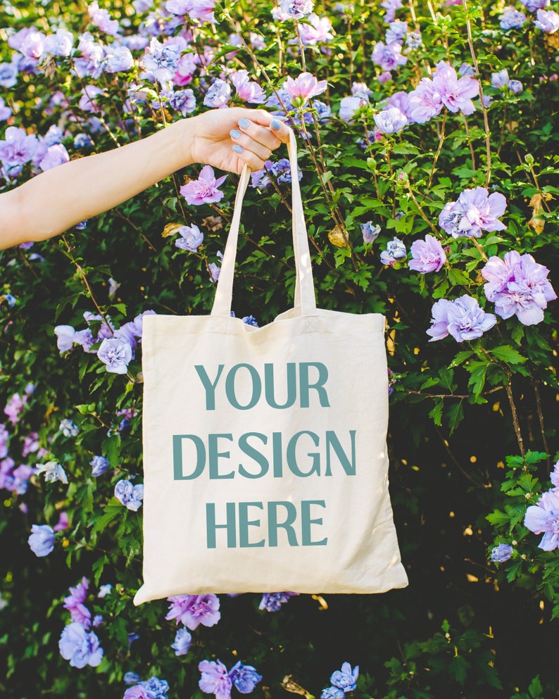 May include: A white canvas tote bag with the text "YOUR DESIGN HERE" printed in teal blue. The bag is held by a hand in front of a bush of purple flowers.