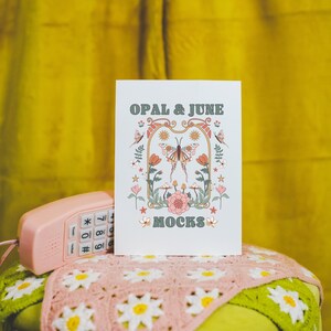 May include: A white card with the text "OPAL & JUNE MOCKS" in green and brown lettering. The card features a colorful illustration of a butterfly with flowers and stars. The card is sitting on a pink crocheted blanket on top of a green stool.