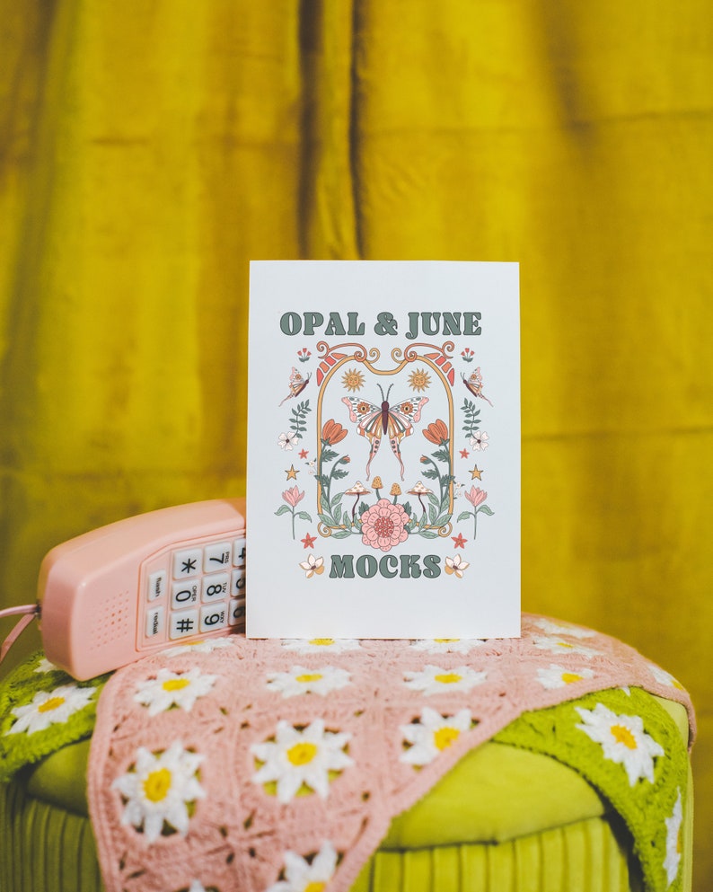 May include: A white card with the text "OPAL & JUNE MOCKS" in green and brown lettering. The card features a colorful illustration of a butterfly with flowers and stars. The card is sitting on a pink crocheted blanket on top of a green stool.