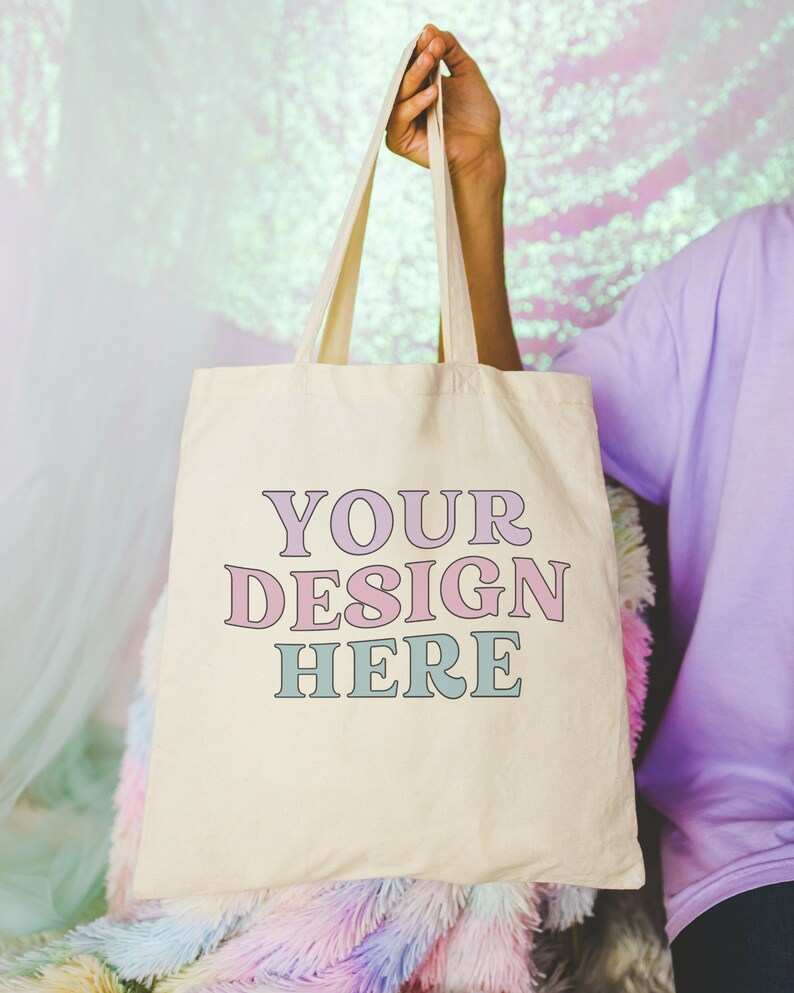 May include: A white canvas tote bag with the text "YOUR DESIGN HERE" in pink and blue lettering.