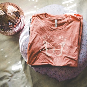 May include: A coral t-shirt featuring the text "OPAL & JUNE" and a butterfly design. The shirt is folded on a round, floral-patterned cushion. A rose gold disco ball is in the upper left corner.