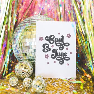 May include: A white card with the text "Opal & June" in black script with pink flowers around the text. The card is sitting on a gold sparkly surface with a silver disco ball and smaller silver disco balls in the foreground.