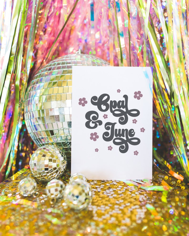 May include: A white card with the text "Opal & June" in black script with pink flowers around the text. The card is sitting on a gold sparkly surface with a silver disco ball and smaller silver disco balls in the foreground.