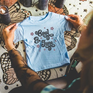 Mockup di maglietta azzurra: foto colorata del modello che piega la maglietta in jersey fine per neonati Light Blue Rabbit Skins 3322 / Whimsigoth Mock per Halloween