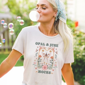 Puede incluir: Una camiseta blanca con un dise&ntilde;o floral y el texto "OPAL & JUNE MOCKS" impreso en la parte delantera. El dise&ntilde;o presenta una mariposa, flores y estrellas.