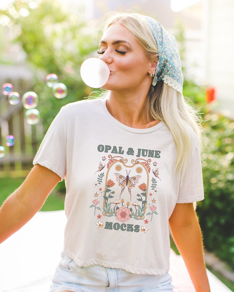 Puede incluir: Una camiseta blanca con un dise&ntilde;o floral y el texto "OPAL & JUNE MOCKS" impreso en la parte delantera. El dise&ntilde;o presenta una mariposa, flores y estrellas.
