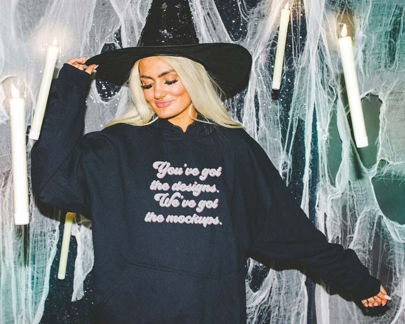 May include: A black hoodie with a pink graphic that says "You've got the designs. We've got the mockups." The model is wearing a black witch hat and is standing in front of a white spiderweb background.