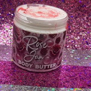 May include: A white jar with a pink lid filled with a white body butter. The jar has a black and pink label with the text "Rose Jam Body Butter" and a small pink rose graphic.