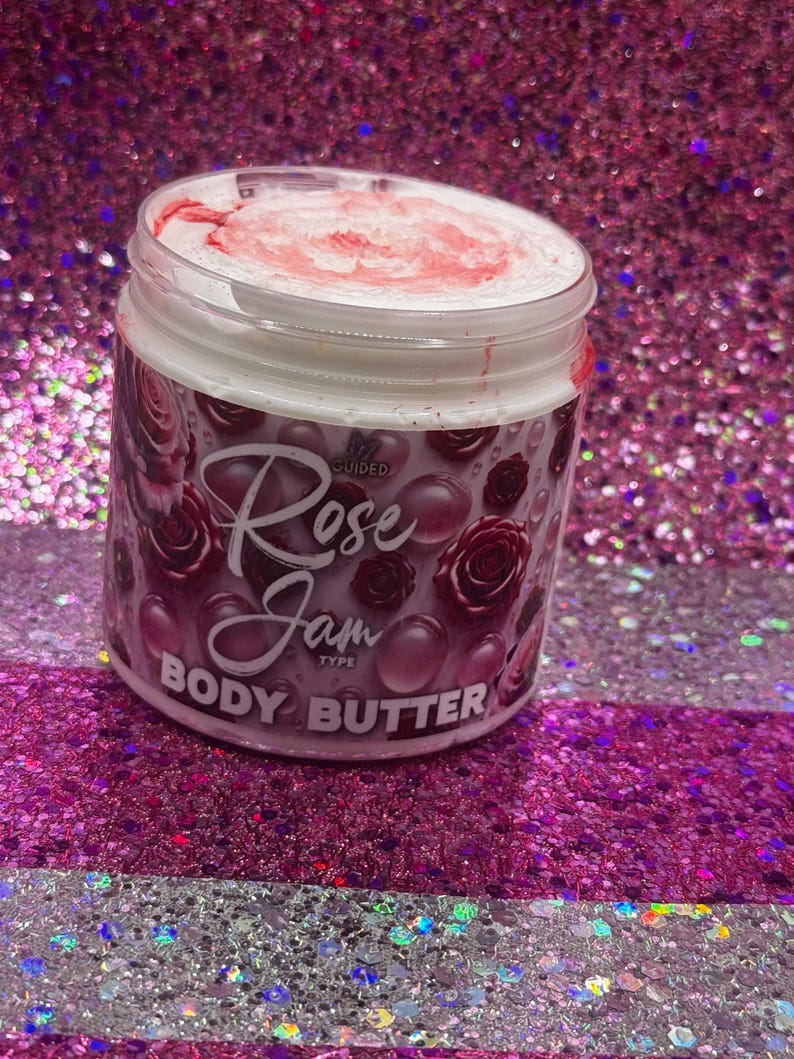 May include: A white jar with a pink lid filled with a white body butter. The jar has a black and pink label with the text "Rose Jam Body Butter" and a small pink rose graphic.