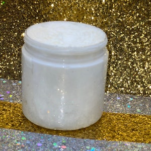 May include: A white jar filled with a white, creamy substance. The jar is sitting on a gold and silver glitter surface.
