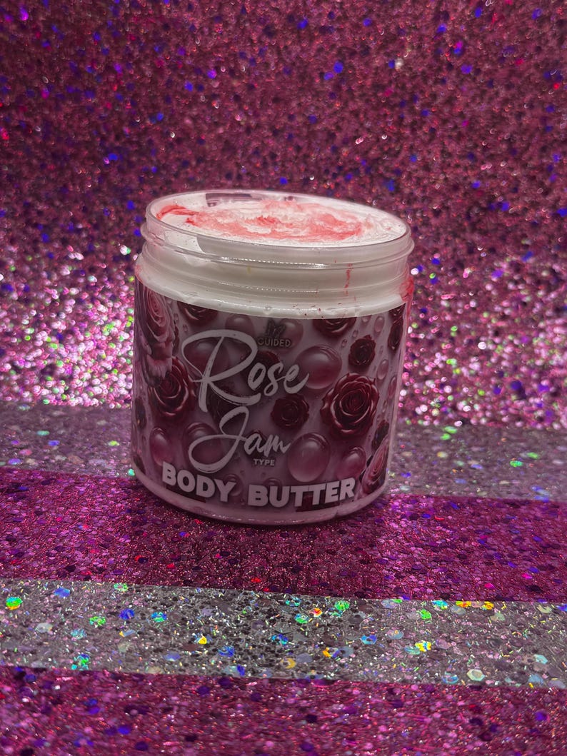 May include: A white jar with a pink lid and a pink and white label that says "Rose Jam Type Body Butter". The jar is sitting on a pink and white glittery surface.