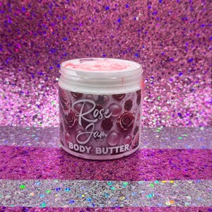 May include: A white jar of body butter with a pink lid. The jar has a label that says "Infused Rose Jam Type Body Butter". The label has a pink and purple floral design.
